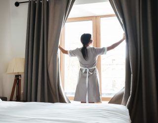 Female housekeeping chambermaid opening window curtains in the hotel room