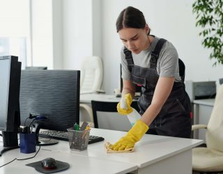 Young female staff of cleaning service company with plastic bottle of detergent wiping desk with duster in openspace office